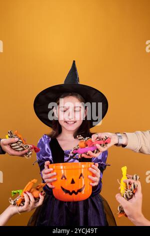 Fille heureuse en chapeau noir et robe de sorcière fantaisie regardant les mains mettant des bonbons et d'autres friandises dans un seau en forme de citrouille Banque D'Images