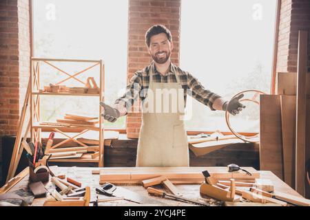 Photo d'homme adulte barbu sympathique portant tablier les mains ouvertes heureux vous voir dans son petit atelier de garage confortable avec des détails en bois Banque D'Images