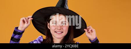 Bannière de mignonne fille souriante en robe de sorcière et chapeau noir debout sur fond orange dans l'isolement et regardant la caméra Banque D'Images