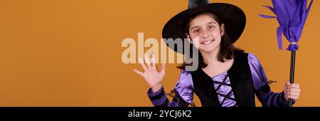 En-tête de fille mignonne dans le costume d'Halloween de sorcière tenant le balai et agitant la main à vous tout en restant debout par copyspace pour votre annonce Banque D'Images