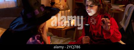 En-tête de garçon mignon en costume de démon d'Halloween griffant et dégourdissant tout en faisant briller son visage par une lampe de poche et racontant l'histoire d'horreur de fille Banque D'Images