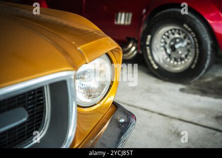 Une vue rapprochée de l'avant d'une voiture jaune classique à côté d'une voiture rouge dans un garage spacieux, mettant l'accent sur le design vintage et le savoir-faire. Banque D'Images