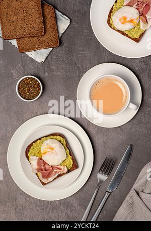 Photographie alimentaire du petit déjeuner nutritionnel, pain de seigle au levain avec de la purée d'avocat, prosciutto, bacon, oeuf poché bouilli, thé avec du lait Banque D'Images