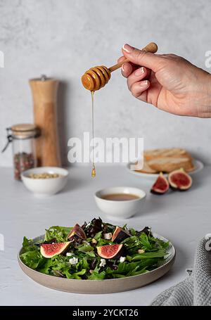 Photographie culinaire de salade à la figue et au fromage feta, épinards, laitue, miel, pignons de pin, pain grillé, table dressée, main verser du miel sur une salade Banque D'Images