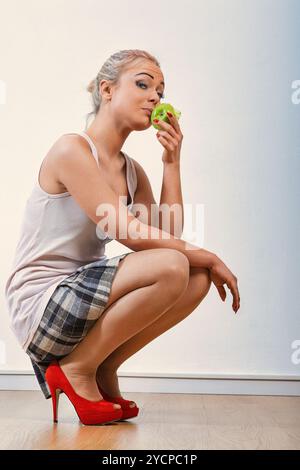 Jeune femme élégante en blanc et plaid. Squatter pour sentir une pomme verte. Exsudant le désir et la beauté avec des talons rouges. Incarner un mode de vie sain. Bouchon Banque D'Images