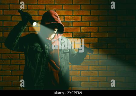 Voleur en sweat à capuche avec pistolet et lampe de poche contre le mur de briques rouges. Vol à main armée Banque D'Images