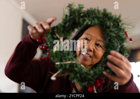 Femme senior asiatique décorant la couronne de Noël à la maison, profitant des préparations festives Banque D'Images
