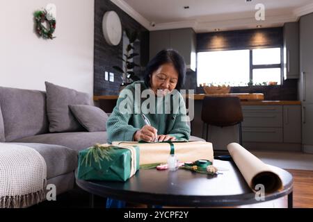 Femme senior asiatique enveloppant cadeau de Noël à la maison, souriant et appréciant l'esprit de vacances Banque D'Images