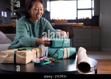 Femme senior asiatique souriante enveloppant des cadeaux de Noël à la maison, profitant des préparations festives Banque D'Images