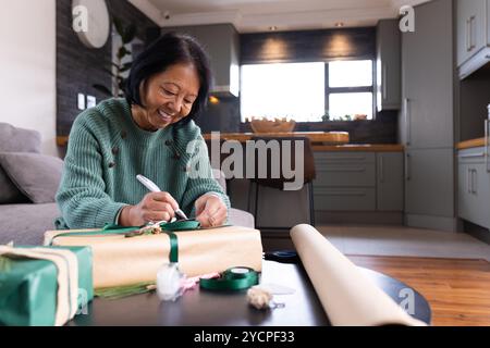 Femme senior asiatique souriante enveloppant des cadeaux de Noël à la maison, profitant des préparations festives Banque D'Images