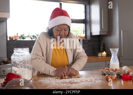 Femme aînée asiatique dans le chapeau de Père Noël faisant des biscuits de Noël à la maison, concentrée et content Banque D'Images