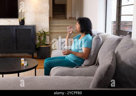 Femme aînée asiatique assise sur le canapé buvant de l'eau, prenant des médicaments à la maison Banque D'Images