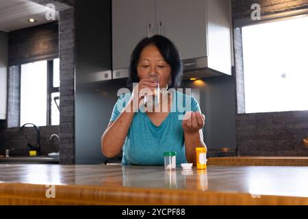 Femme aînée asiatique prenant des médicaments avec de l'eau dans la cuisine moderne à la maison Banque D'Images