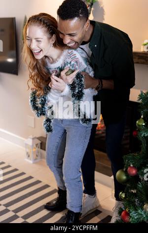 Décorer l'arbre de Noël avec guirlande, couple multiracial riant ensemble à la maison Banque D'Images