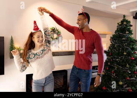 Heureux couple multiracial dansant joyeusement près du sapin de Noël à la maison, portant des pulls festifs Banque D'Images