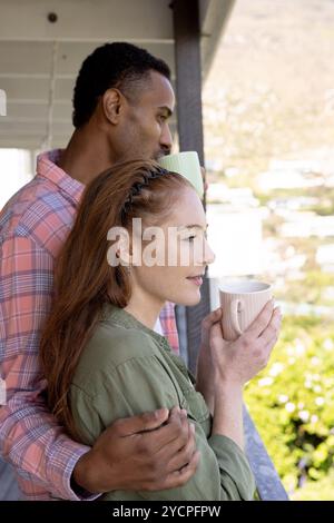 Couple multiracial profitant du café du matin sur le balcon, embrassant et souriant paisiblement Banque D'Images