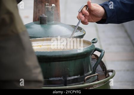 Vue d'un pot en métal vert avec un couvercle ouvert et une louche, rempli de soupe chaude sur une installation rustique de cuisine extérieure. Banque D'Images