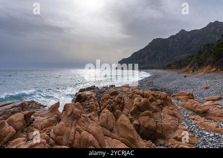 Côte sauvage et accidentée de Sardaigne avec des vagues qui s'écrasent sur la plage rocheuse et des rochers de granit rouge au premier plan Banque D'Images