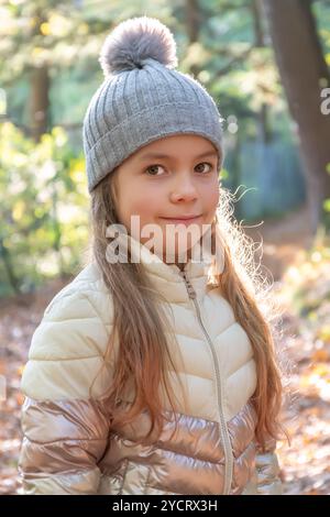 Portrait d'une petite fille aux cheveux longs vêtue d'une veste et d'un chapeau gris, debout tranquillement dans une forêt ensoleillée entourée de feuilles d'automne brillantes. Banque D'Images