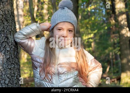 Une jeune fille joyeuse se tient dans la rue, portant un bonnet tricoté et une veste en duvet brillante. Les feuilles d'automne colorées créent un beau fond. Banque D'Images