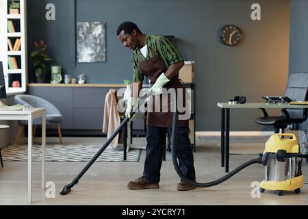 Vue latérale pleine longueur d'un jeune homme afro-américain travaillant dans le service de nettoyage et aspirant les planchers dans la maison ou le bureau, espace de copie Banque D'Images
