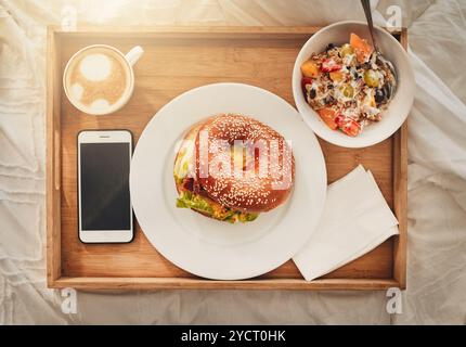 Bagel, plateau et nourriture avec café au-dessus sur le lit pour le service en chambre, repas ou petit déjeuner à l'hôtel. Vue de dessus, pain et collation avec écran de téléphone, cappuccino ou Banque D'Images