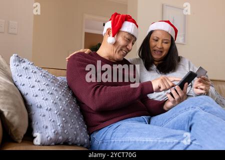 Noël, couple multiracial senior portant des chapeaux de Père Noël magasinant en ligne avec smartphone à la maison Banque D'Images