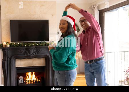 Noël, couple multiracial senior dansant joyeusement près de la cheminée, portant des chapeaux de Père Noël à la maison Banque D'Images