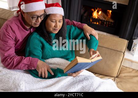 Temps de Noël, couple multiracial senior dans le livre de lecture de chapeaux de Père Noël par cheminée confortable à la maison Banque D'Images