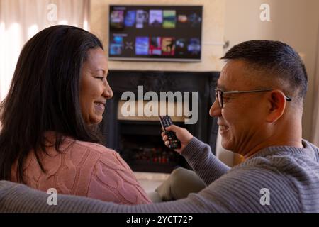 Couple senior multiracial profitant d'une soirée cinéma confortable à la télévision à la maison, souriant et se relaxant ensemble Banque D'Images