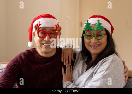 Noël, couple multiracial senior portant des chapeaux et des lunettes de fête, souriant joyeusement à la maison Banque D'Images