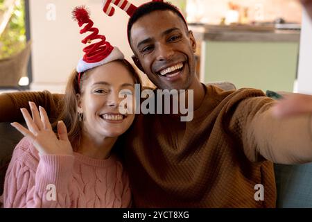 Noël, couple multiracial portant des chapeaux festifs prenant selfie, souriant joyeusement à la maison Banque D'Images