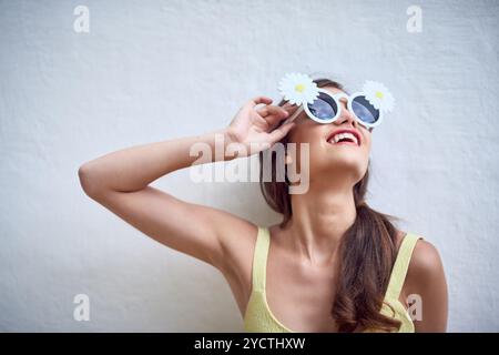 Mur, femme et fleurs dans des lunettes de soleil pour la mode, style tendance et sourire avec pose pour la fierté. Personne féminine, confiance et Marguerite avec accessoire pour Banque D'Images