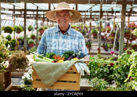 Homme âgé souriant tout en tenant des légumes fraîchement récoltés dans une caisse en bois, portant un chapeau de paille et debout dans une serre luxuriante remplie de plantes et de fleurs vibrantes Banque D'Images