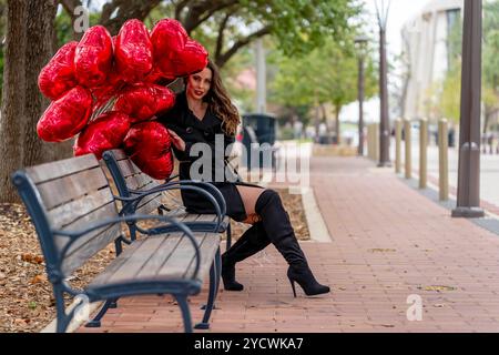 Une magnifique brune se trouve sur un banc de parc, son sourire brillant alors qu'elle attend son amant. Serrant une douzaine de ballons rouges en forme de cœur, elle rayonne d'amour Banque D'Images