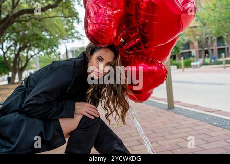 Une magnifique brune se trouve sur un banc de parc, son sourire brillant alors qu'elle attend son amant. Serrant une douzaine de ballons rouges en forme de cœur, elle rayonne d'amour Banque D'Images