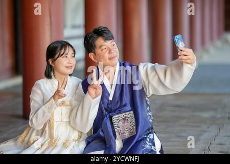 Un couple coréen, un homme dans ses 30 ans et une femme dans ses 20 ans, portent joyeusement Hanbok et prennent un selfie avec un smartphone à l'intérieur d'un bâtiment historique Banque D'Images