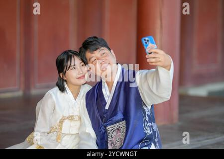 Un couple coréen, un homme dans ses 30 ans et une femme dans ses 20 ans, portent joyeusement Hanbok et prennent un selfie avec un smartphone à l'intérieur d'un bâtiment historique Banque D'Images