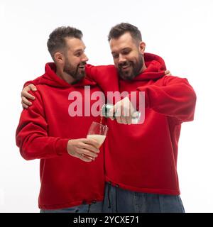 Portrait de deux jeunes frères vêtus de tenues assorties posant ensemble, quand un frère verse de la bière au verre dans les mains d'un autre contre blanc Banque D'Images