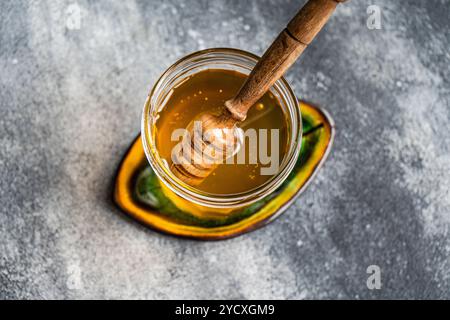 Vue de dessus de miel biologique doré dans un bocal en verre transparent, avec une trempette à miel en bois partiellement immergée. Capturé sur une surface grise texturée avec un Banque D'Images