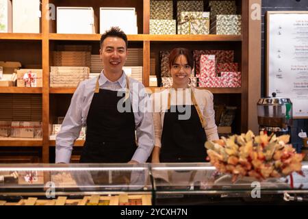 Deux employés joyeux dans des tabliers debout derrière un comptoir dans un magasin de chocolat bien approvisionné, souriant vers la caméra Banque D'Images