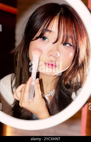 Une jeune femme asiatique applique le blush à l'aide d'un pinceau de maquillage, regardant attentivement son reflet dans un miroir circulaire, soulignant son attention aux détails Banque D'Images
