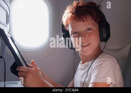 Un jeune garçon joyeux portant des écouteurs regarde directement la caméra tout en tenant une tablette à bord d'un avion très éclairé. Son expression joyeuse ajoute Banque D'Images