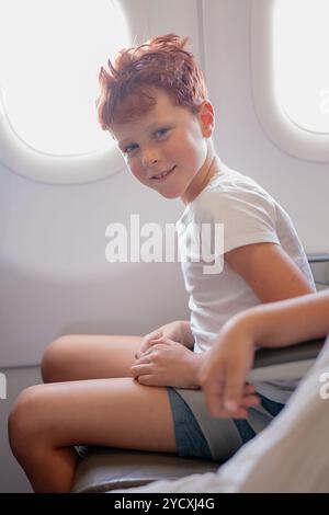 Vue de côté d'un garçon préadolescent aux cheveux roux, portant un t-shirt blanc, assis confortablement sur un siège d'avion près de la fenêtre, sourit en regardant directement Banque D'Images