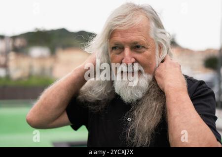 Un homme âgé avec une barbe grise coulante et de longs cheveux tient ses cheveux, envisageant un don pour soutenir les patients atteints de cancer qui ont perdu leurs cheveux Banque D'Images