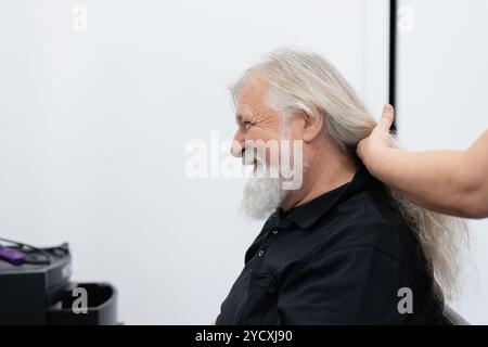 Un vieil homme joyeux avec une barbe grise et de longs cheveux sourit tout en se faisant préparer pour don par une personne derrière lui Banque D'Images