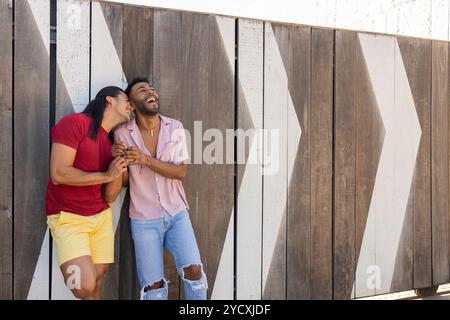 Un couple joyeux et multiethnique gay partage un rire sur un fond géométrique moderne, soulignant leur affection et l'environnement urbain dynamique Banque D'Images