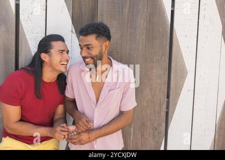 Un couple joyeux et multiethnique gay partage un rire sur un fond géométrique moderne, soulignant leur affection et l'environnement urbain dynamique Banque D'Images