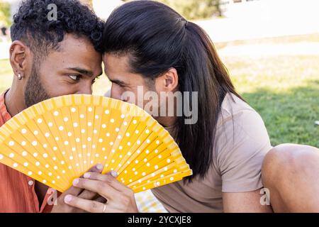 Un moment affectueux entre un couple gay interracial, partageant des murmures derrière un fan jaune vif dans un parc de la ville, symbolisant l’amour et le PRI LGBTQIA+ Banque D'Images