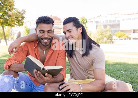 Un joyeux couple gay, mettant en valeur un mélange latino-américain et afro-américain, se détend dans un parc urbain qu’ils partagent un moment proche tandis que l’on lit un livre A. Banque D'Images